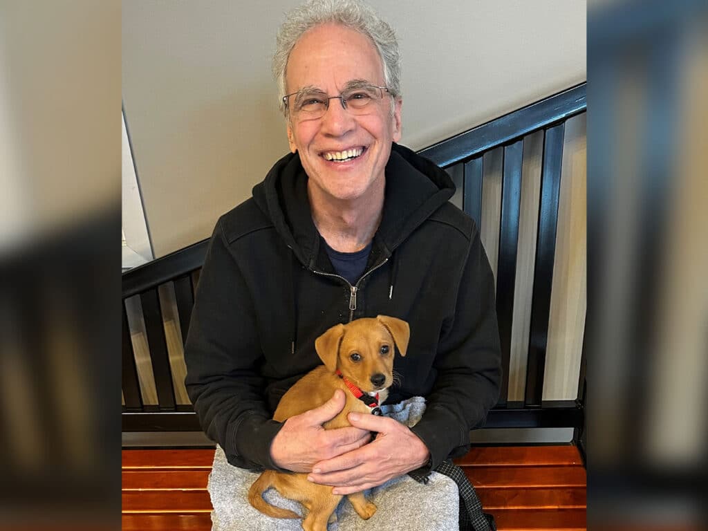 A smiling man with gray hair and glasses sits on a bench, holding a small brown puppy with a red collar. The puppy is nestled comfortably in his lap, and the background features a wooden slatted wall.