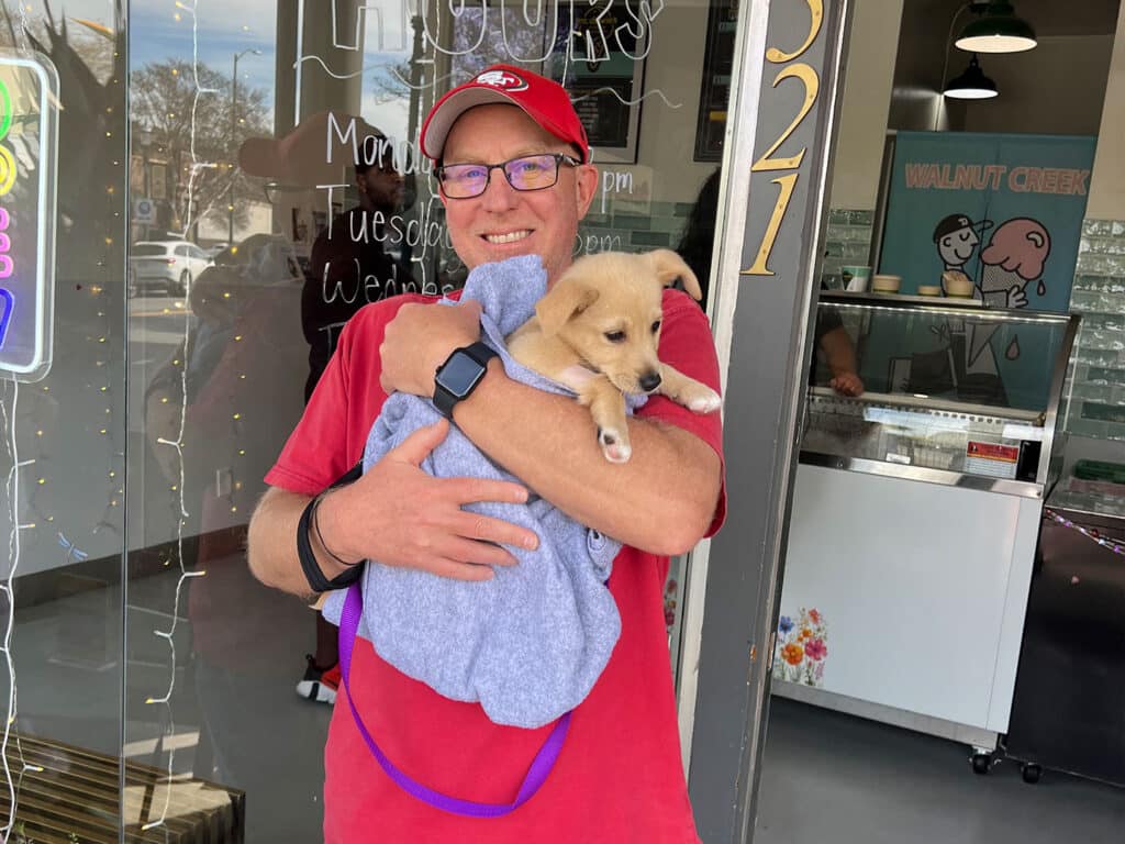A man wearing glasses and a red cap smiles while holding a small puppy wrapped in a light blue blanket outside a bright shop. The store's name is partially visible in the background, along with festive lights.