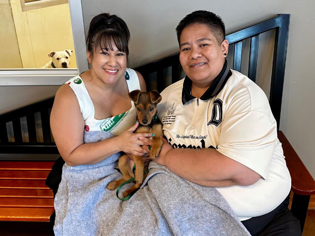 A woman and a person sitting on a bench, both smiling while holding a small brown puppy. The woman is in a white dress with green accents, and the person is wearing a black and white sports shirt. A dog can be seen in the background.