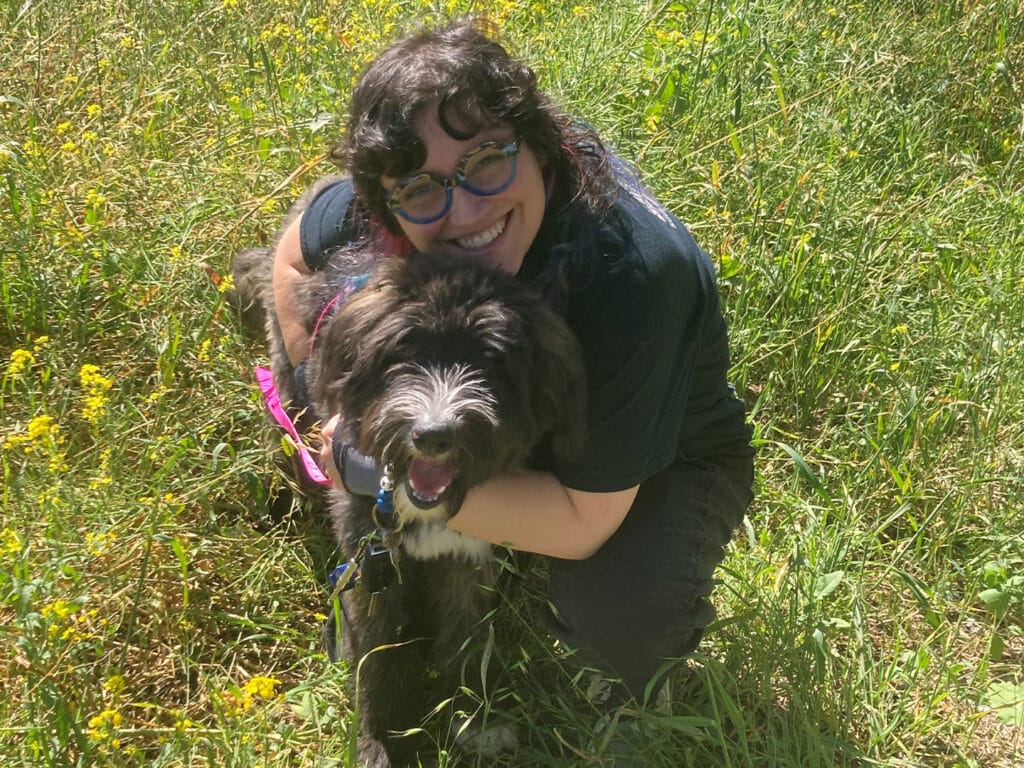 A woman with curly hair and glasses kneels in a grassy field, smiling while hugging a medium-sized dog. The dog has a fluffy coat and is wearing a harness. Yellow wildflowers are visible in the background.