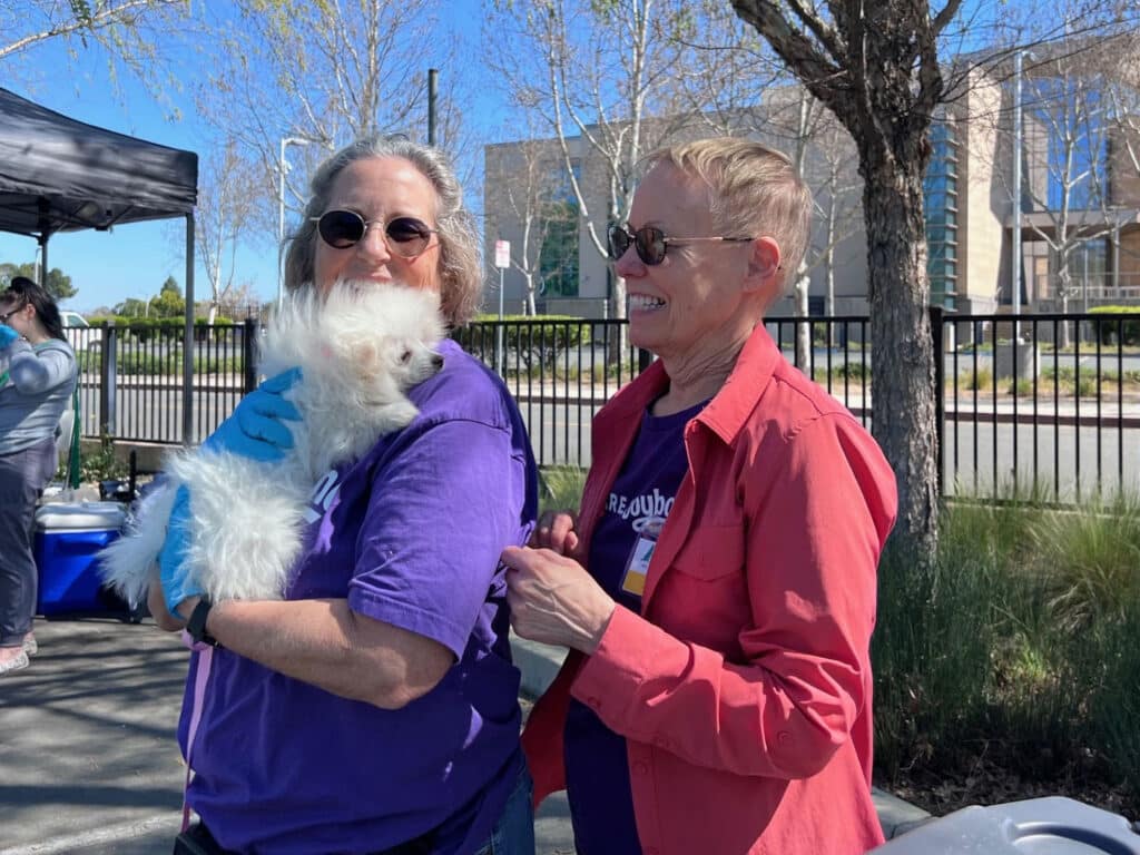 Two women stand outdoors at a community event. One woman, wearing glasses and a purple shirt, holds a small white dog. The other woman, wearing a red jacket, smiles and interacts with her. A table and trees are visible in the background.