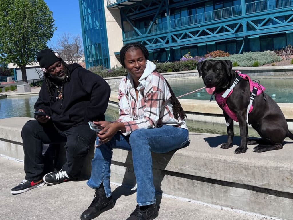 Two individuals sitting on a concrete ledge near a fountain. On the left, a man with a beard in a black hoodie and sneakers uses a mobile device. Next to him is a woman wearing a plaid jacket and blue jeans. A black dog in a pink harness sits beside them.