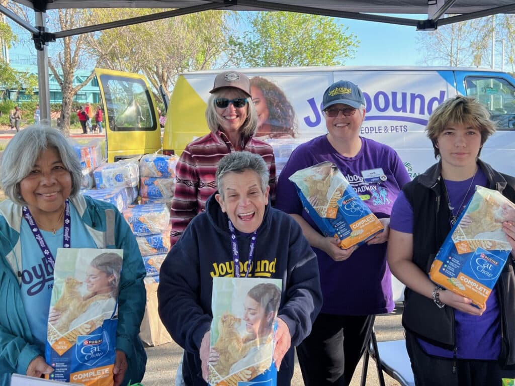 Five individuals stand under a tent at a community event, each holding bags of pet food. They are smiling and wearing matching volunteer shirts. In the background, a van branded with 'Joyound' is parked, surrounded by more pet food bags.