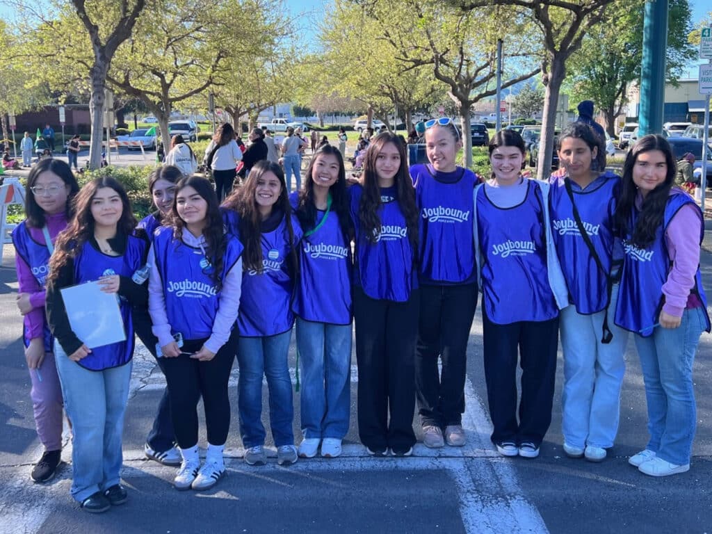 A group of ten volunteers wearing purple vests stands together in a parking lot, smiling at the camera. Trees are visible in the background, and other people can be seen participating in an event.