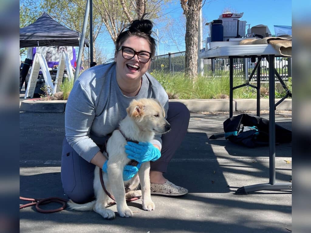 A woman in a gray shirt and blue gloves crouches on a paved area, smiling while holding a small golden puppy. In the background, there are trees and a canopy structure. The scene appears to be outdoors, possibly at a community event.