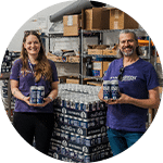 Two individuals stand in a warehouse setting, each holding a container of product. The man has long hair and a beard, while the woman is smiling. Behind them are shelves filled with various items and stacks of products.