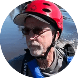 A man wearing a red helmet and sunglasses, with gray hair and a beard, is seen against a body of water. He is dressed in a blue life jacket, suggesting he is in an aquatic or outdoor environment.