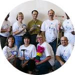 A group of nine people in white t-shirts with a logo, posing together in a well-lit room. They hold various objects, including paint brushes and colorful decorations, indicating a community or team event. The backdrop is plain.