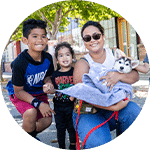 A smiling woman wearing sunglasses holds a small dog wrapped in a blanket while standing next to two children. One child is smiling broadly, and the other is looking at the camera. The background features trees and buildings, suggesting an outdoor event.