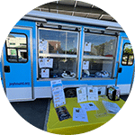 A blue food truck with multiple windows, showcasing various printed materials and brochures displayed on a yellow table. The surrounding area is open, with daylight visible and trees in the background.