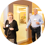 A woman and a man entering a room, the woman holds a drink and wears a black top with a patterned skirt. The man, dressed in a blue and white striped shirt, walks beside her. The setting has a warm atmosphere with light-colored walls.