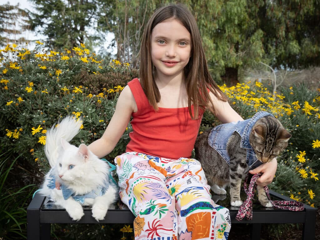 A young girl in a red shirt sits on a bench surrounded by yellow flowers, holding a white cat in a denim jacket on her left and a gray tabby cat in a denim vest and pink leash on her right, smiling at the camera.