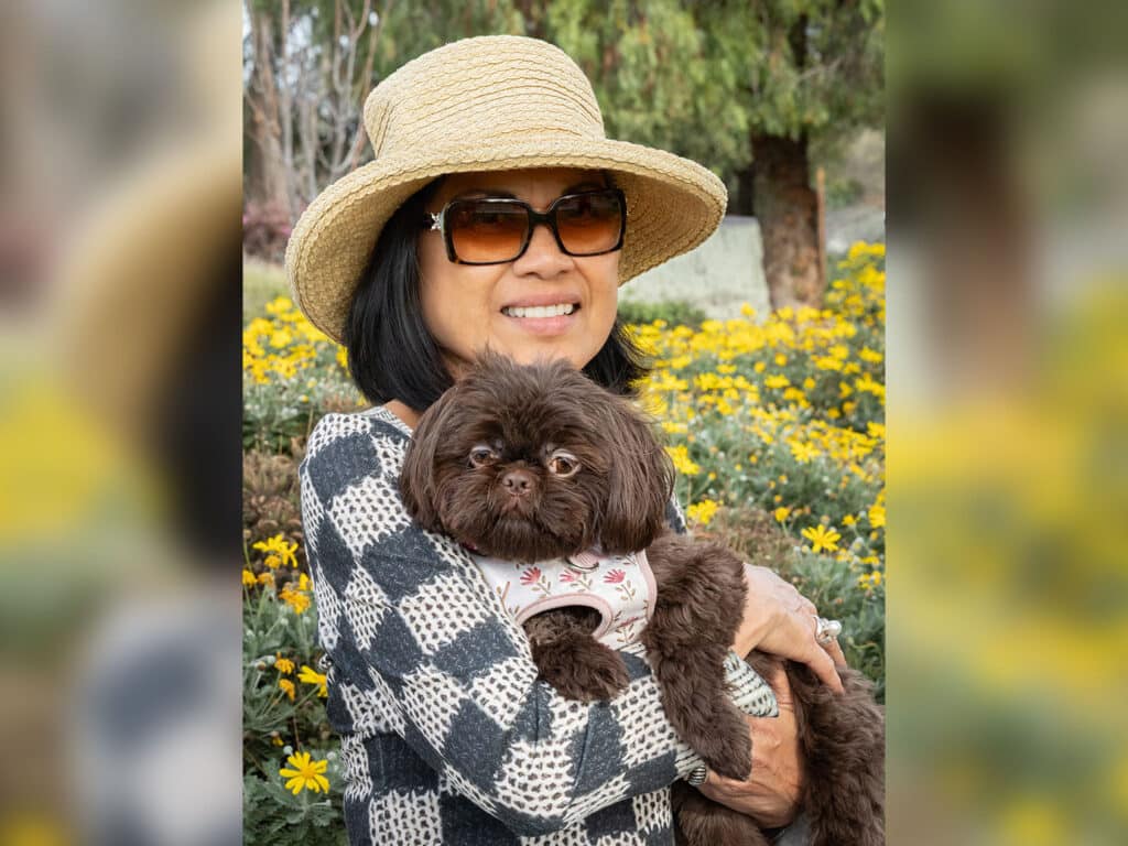 A woman wearing a wide-brimmed straw hat and sunglasses holds a small brown dog in a floral harness. They are surrounded by blooming yellow flowers in a garden setting.