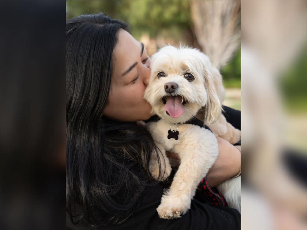 A woman holds a small, fluffy dog close to her face while she kisses its head. The dog has a white coat and appears happy with its mouth open. The background features greenery, suggesting a sunny outdoor setting.