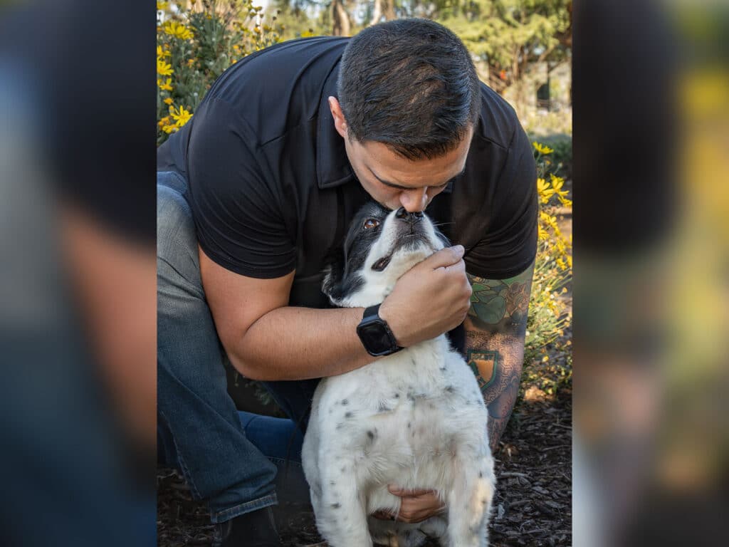 A person kneeling in a garden, leaning in to touch noses with a black and white dog. The person has short hair, wearing a black shirt, while the dog sits on the ground with its mouth slightly open. Surrounding greenery is visible.