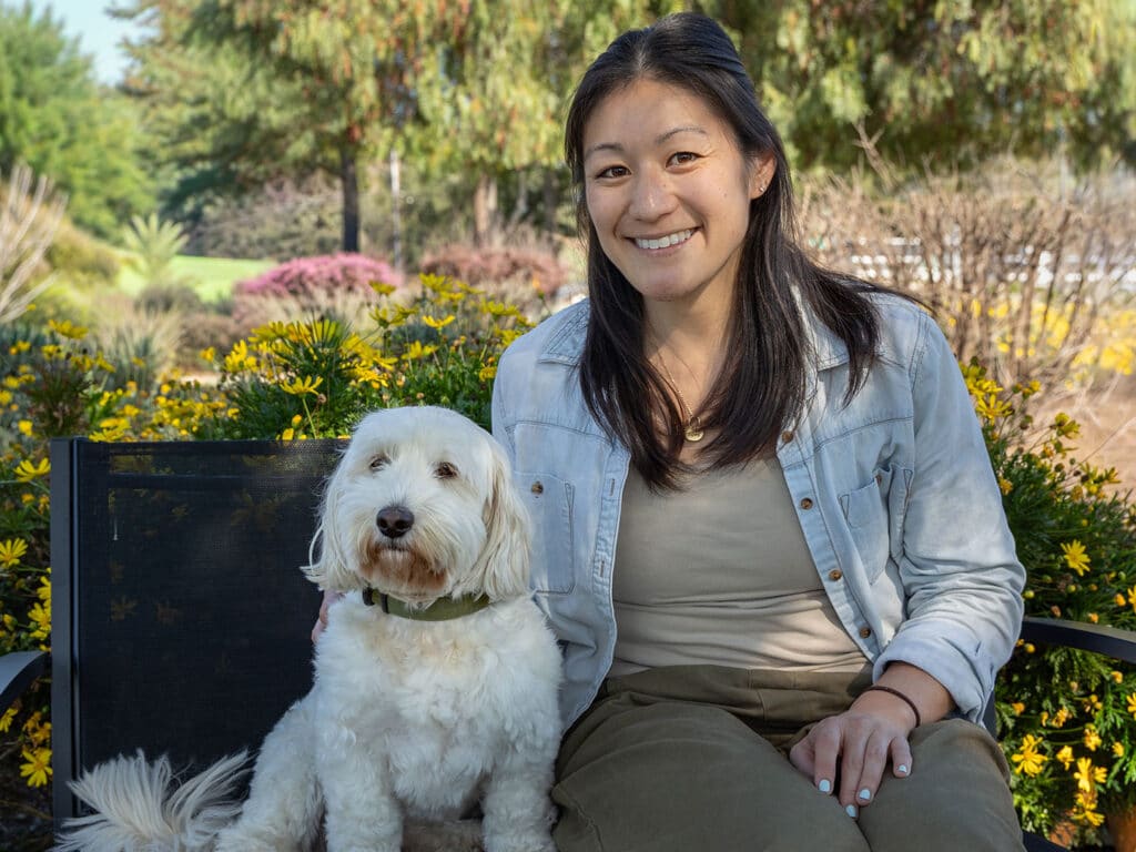 A woman with long dark hair sits on a bench next to a white dog in a green collar. They are surrounded by bright yellow flowers and greenery in a garden setting, capturing a sunny spring day.