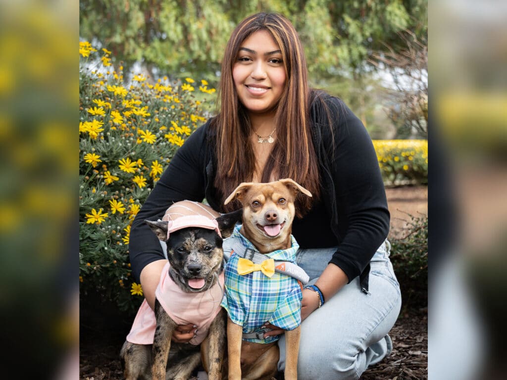 A young woman sitting outdoors with two dogs. One dog is wearing a pink outfit with a hat, and the other is dressed in a blue plaid shirt with a yellow bow tie. They are surrounded by lush greenery and yellow flowers.
