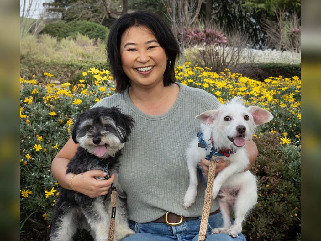 A woman with dark hair smiles while holding two small dogs. One dog is black with gray fur and the other is white with light brown spots. They are surrounded by blooming yellow flowers in a garden.