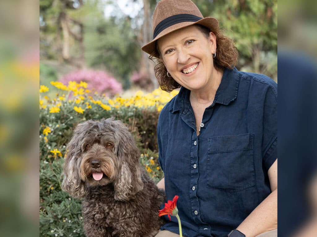 A woman wearing a blue shirt and a brown hat smiles while sitting next to a brown dog in a lush garden filled with yellow flowers. The dog has a playful expression, and the scene captures a springtime outdoor ambiance.