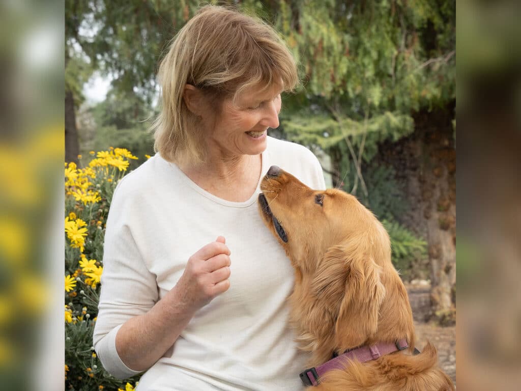 A woman in a white long-sleeve shirt smiles at a golden retriever standing beside her. They are outdoors among yellow flowers, with blurred greenery in the background, creating a relaxed spring atmosphere.