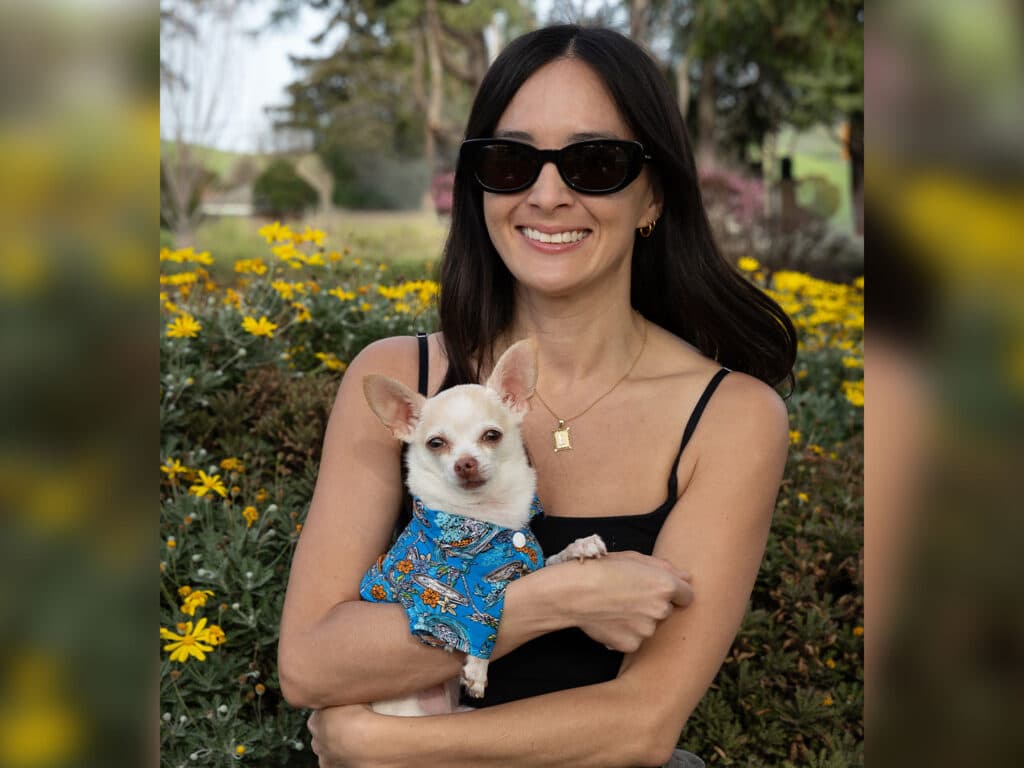 A woman wearing sunglasses smiles while holding a small, light-colored dog in her arms. The dog is dressed in a blue patterned outfit. They are surrounded by yellow flowers and greenery in a springtime setting.