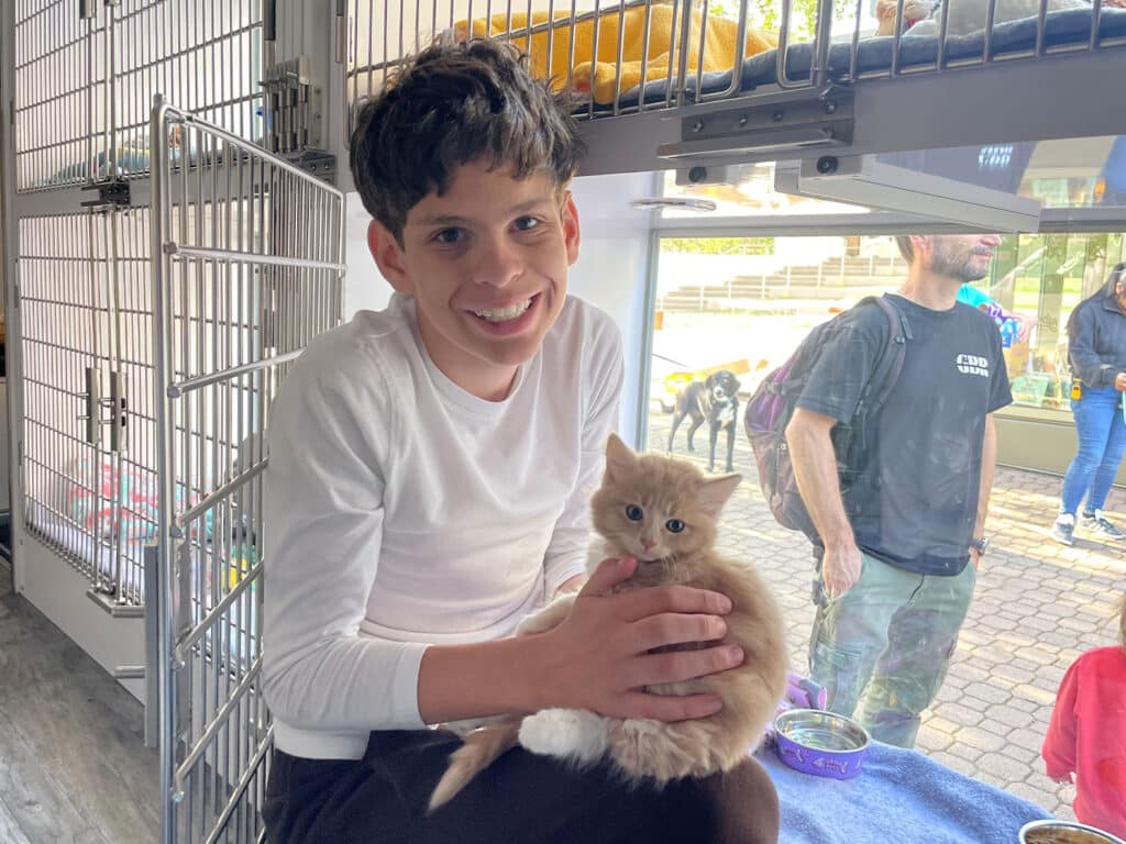 A smiling boy with short dark hair holds a fluffy light brown kitten in his lap, sitting on a bench in a pet shelter. Behind them, there are two adults and various cat habitats visible.