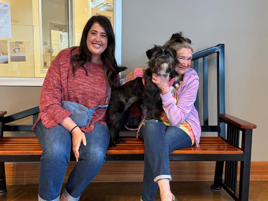 A woman wearing a red sweater and another woman in a purple hoodie sit on a bench in a well-lit room. They are smiling while holding a small, dark-colored dog between them, showcasing a joyful moment together.