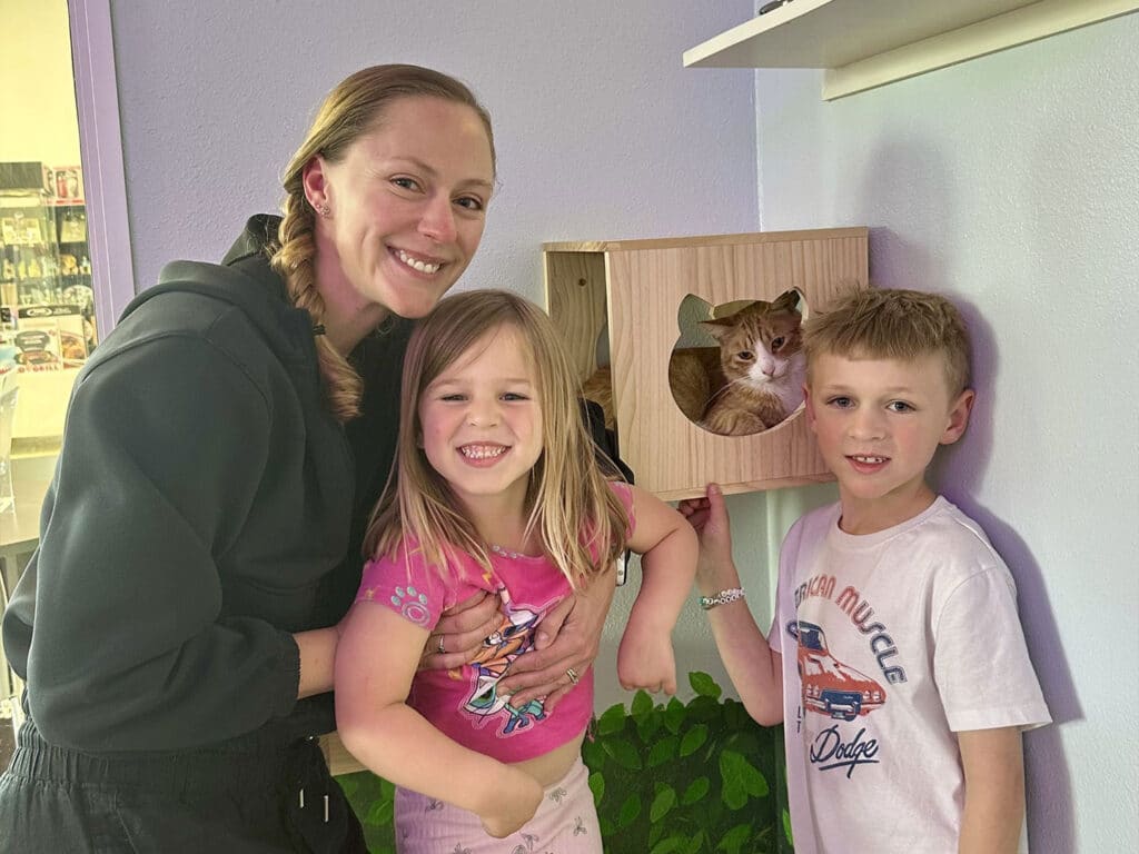 A woman stands with two children in a pet adoption center. The woman holds one child, while the other child stands next to a wooden cat house. A cat is visible inside the house, and the walls are painted in soft colors.