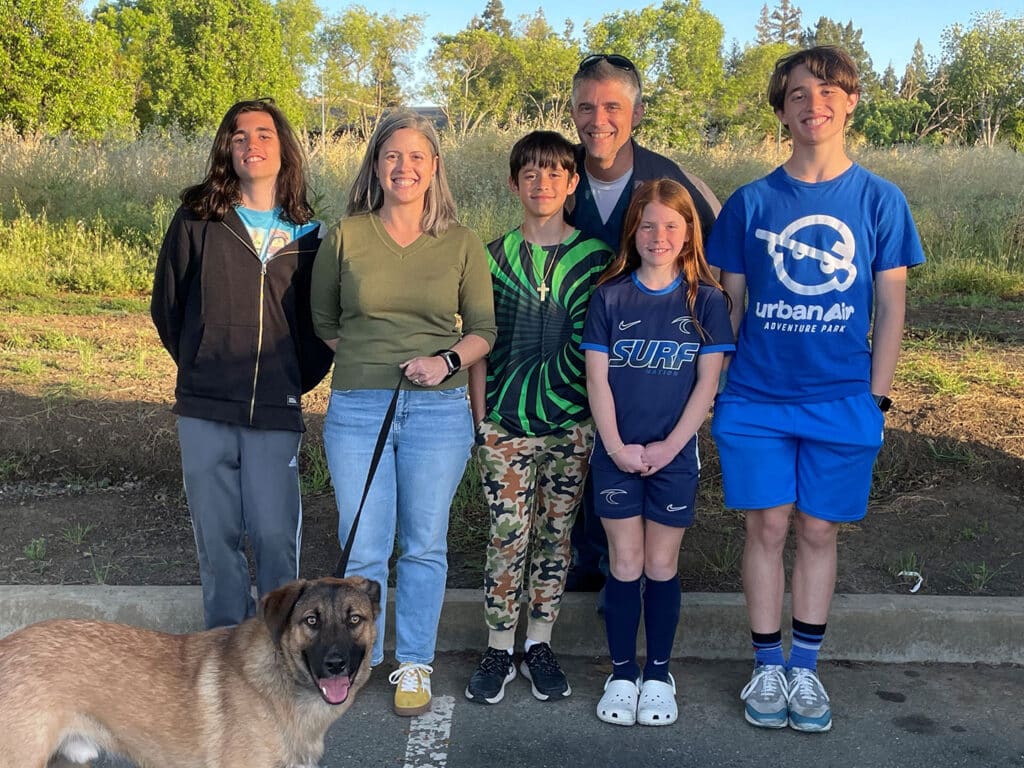 A family poses together outdoors, smiling. The group includes four children and two adults, with one adult holding a dog on a leash. They are in a park-like setting with trees in the background and standing on a paved surface.