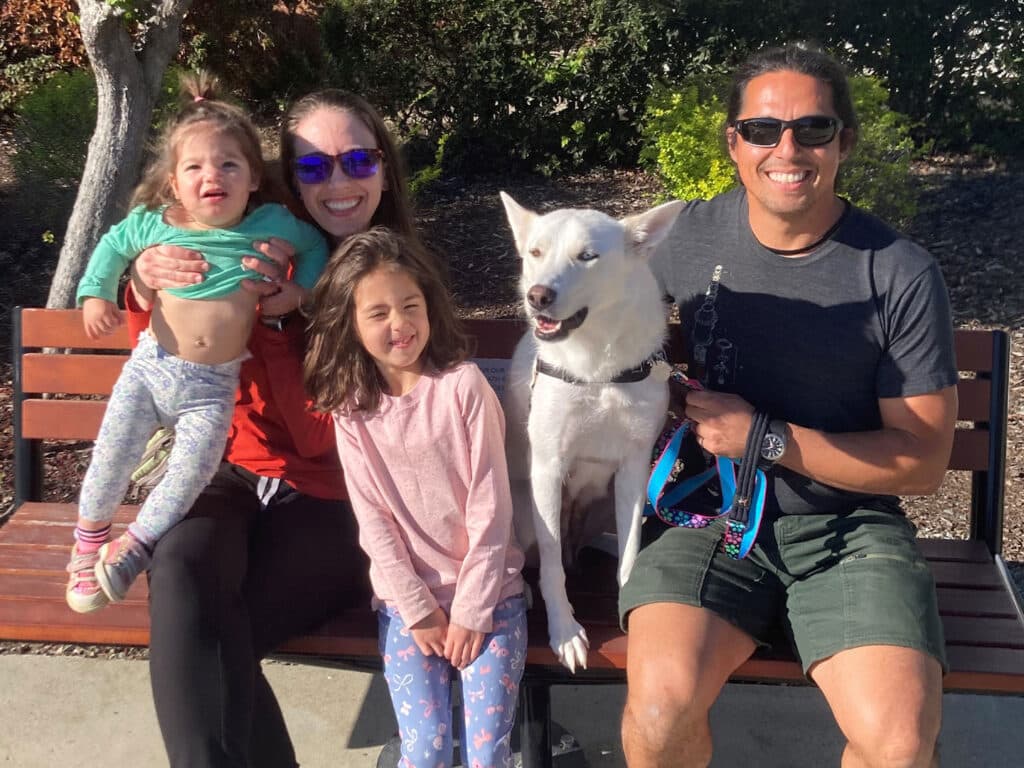 A family of four sits on a wooden bench in a park. They include two young girls, one woman holding a toddler, and a man beside a white dog. The scene features leafy trees and shrubs in the background, suggesting a sunny day.