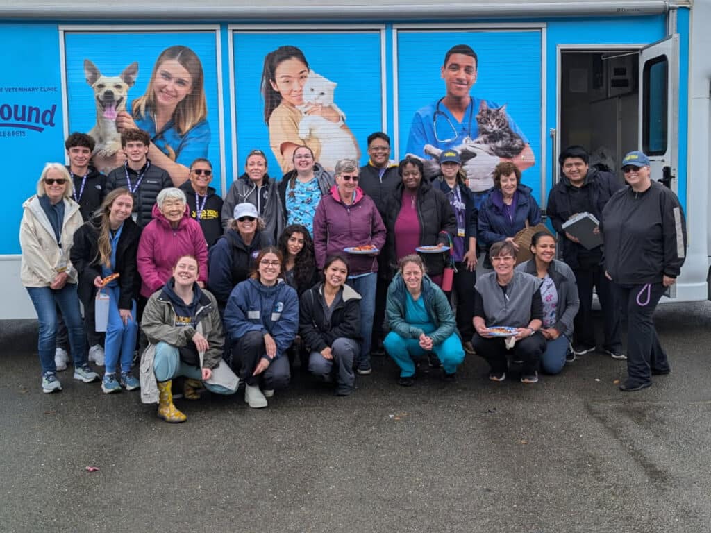 A diverse group of 22 people gathered outdoors in front of a vehicle, smiling and posing for a photo. The background features a promotional display with images of pets and people. Participants wear various outfits, including jackets and scrubs.