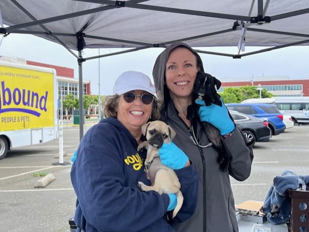 A woman in a blue jacket and a cap holds a puppy, smiling at the camera. Next to her, a veterinarian in a gray coat cradles another puppy, wearing blue gloves. A food truck and parked cars are visible in the background under a tent.