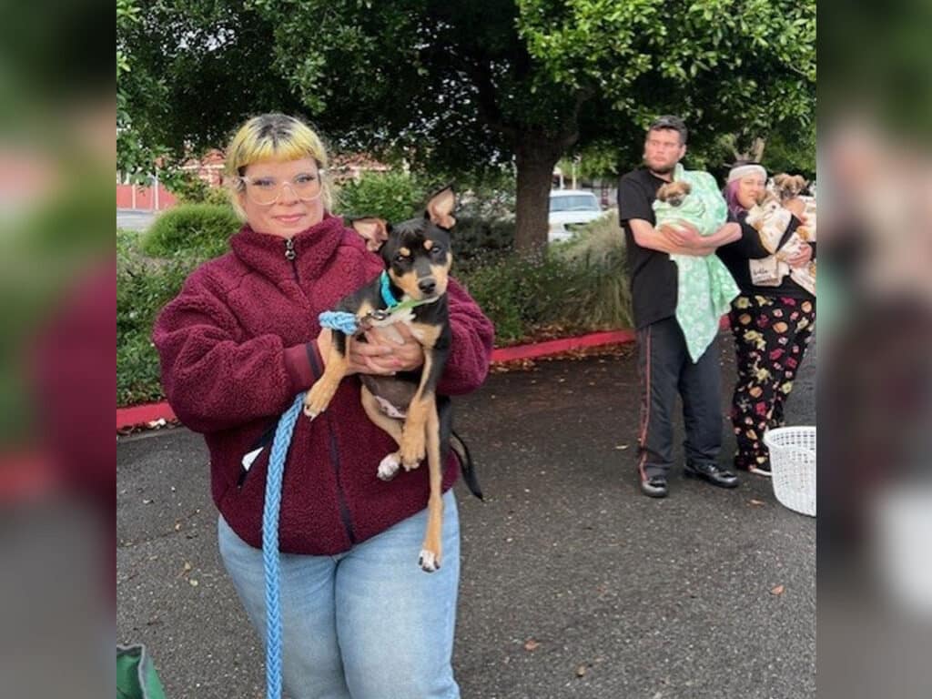 A woman wearing a red fleece jacket holds a small brown dog with a blue collar, standing outdoors. In the background, two people hold animals, and a basket is visible on the ground. Trees are present in the surrounding area.