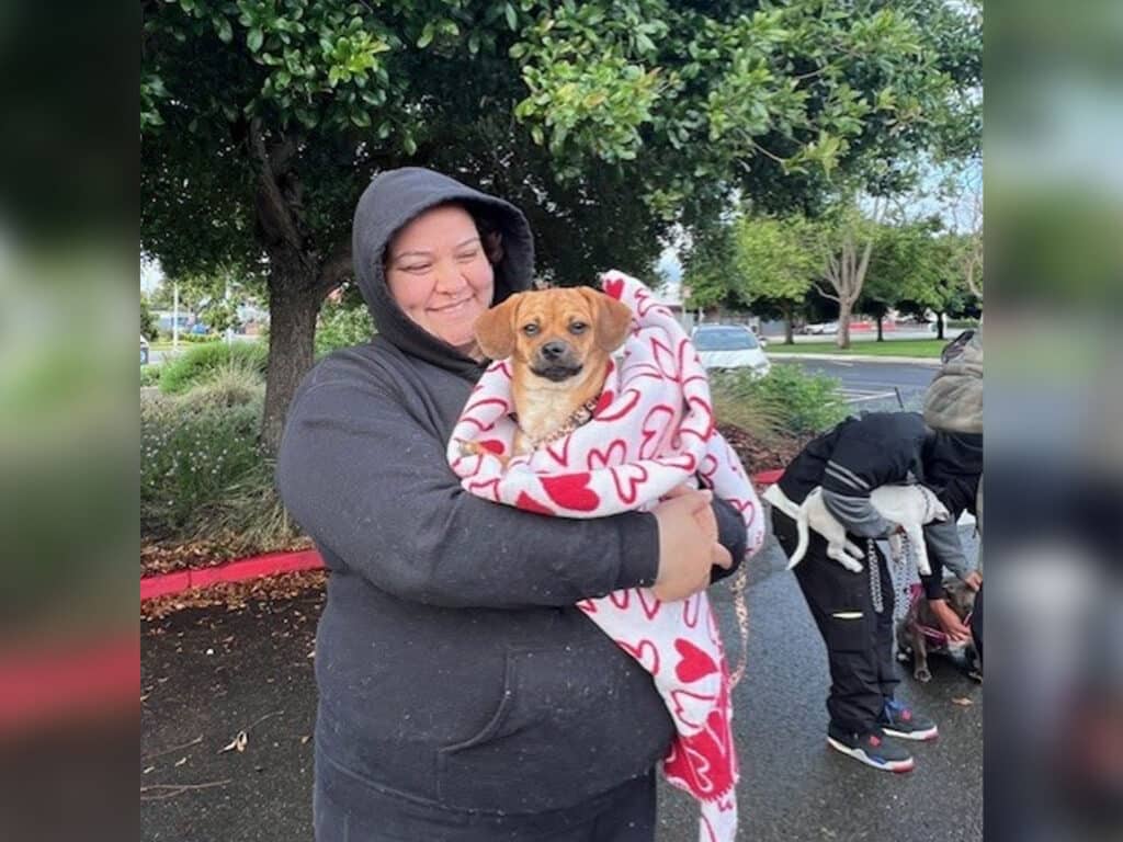 A woman holding a small dog wrapped in a blanket while standing outdoors. The woman is wearing a black hoodie and appears to be smiling. In the background, other people and dogs can be seen. Trees are visible in the surrounding area.