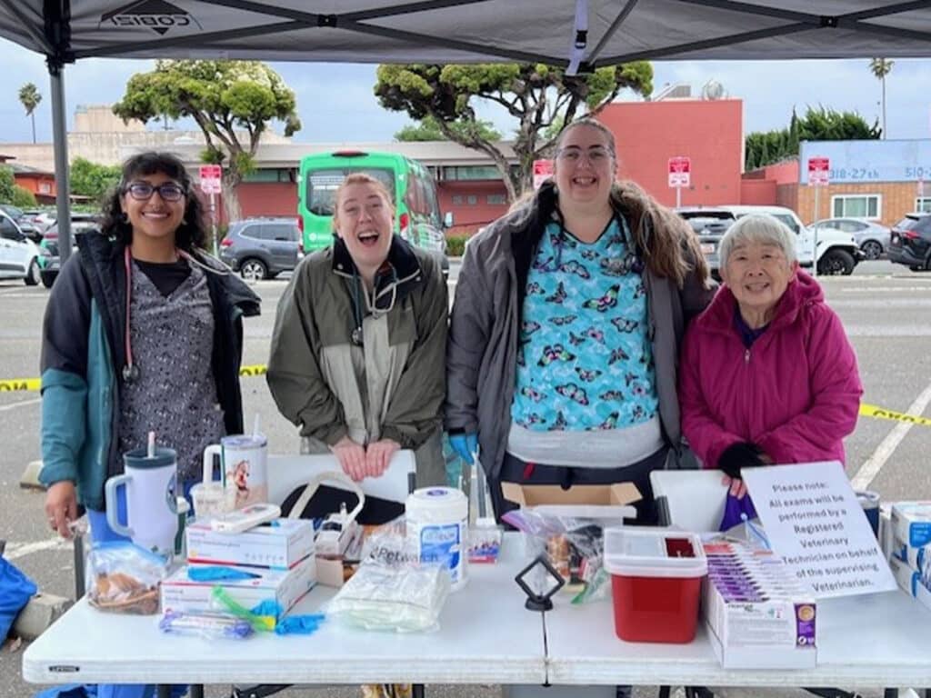 Four volunteers stand behind a table full of medical supplies and informational materials at a community event. They are smiling and interacting with each other. In the background, there are vehicles and trees.
