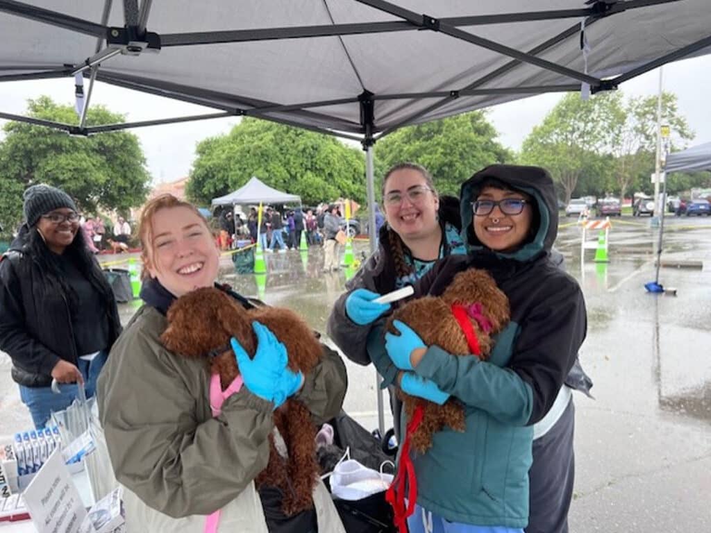 Three women smile while holding small brown puppies under a tent during a rainy outdoor event. The women wear gloves, and there are medical supplies visible on a table nearby. In the background, additional tents and people are seen.