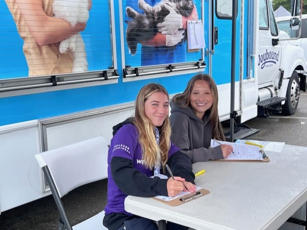 Two young women sitting at a table in front of a truck, each writing on a clipboard. The background features promotional graphics of pets. One woman has long, wavy hair and wears a purple shirt, while the other has straight hair and wears a gray hoodie.