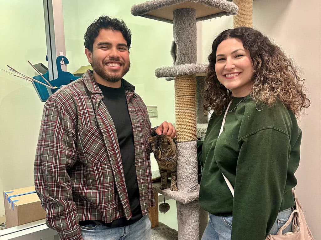 A man and a woman smile while standing in front of a cat tree in a pet adoption center. The man wears a plaid shirt over a dark top, and the woman is dressed in a green sweatshirt. A cat is near the woman's hand.