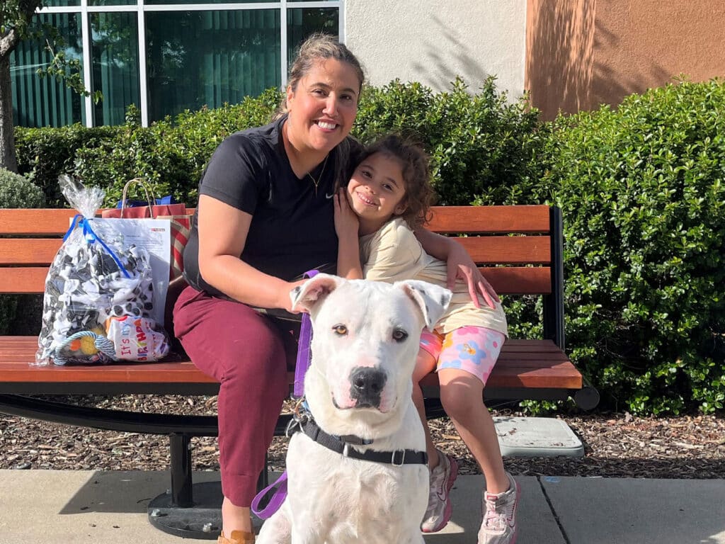 A woman and a young girl sit on a wooden bench outside. The woman, with long hair and wearing a black shirt, smiles while hugging the girl. The girl, dressed in light-colored clothes, leans against her. A large white dog sits in front of them, and a bag of items is placed next to the bench.