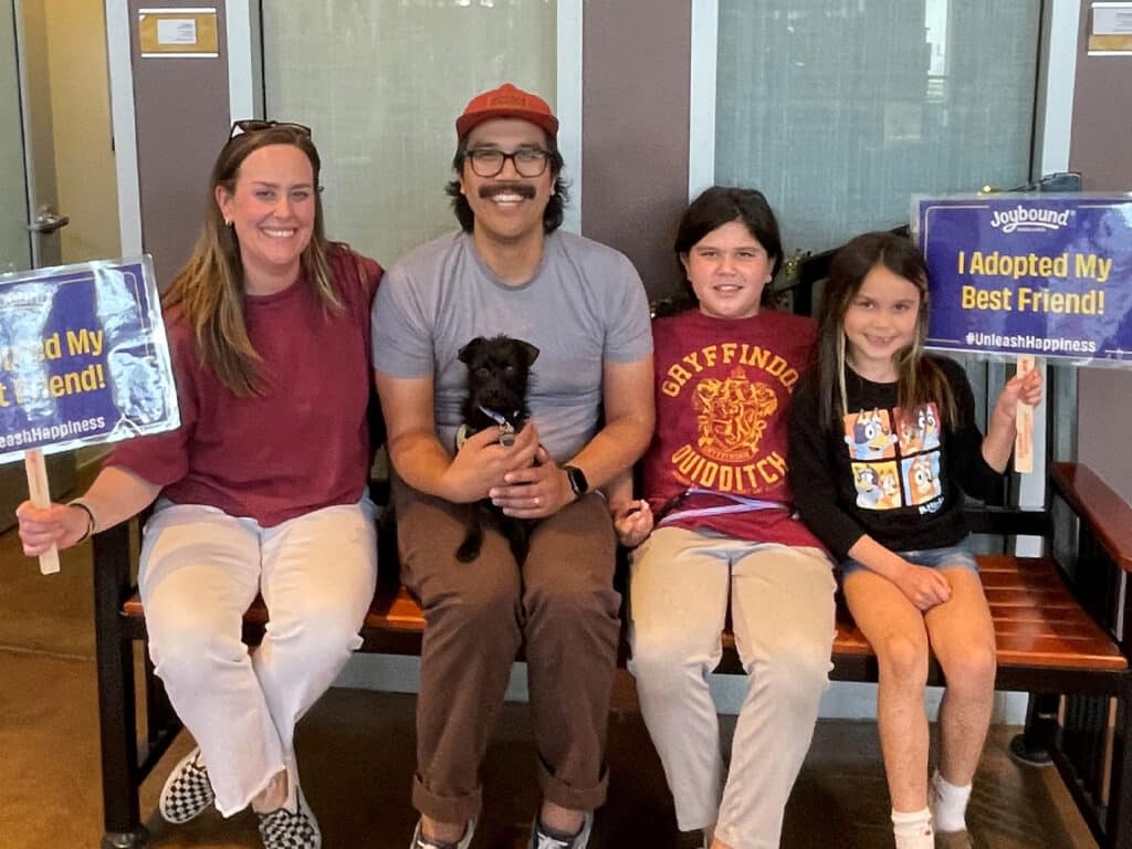 A man wearing glasses and a red hat sits on a bench, holding a small black dog. He is flanked by a woman and two children, each holding signs that say "I Adopted My Best Friend!" and featuring the hashtag #UnleashHappiness.