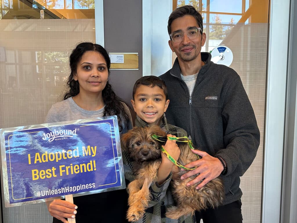 A woman and a man stand with a boy holding a small dog, all smiling. The woman holds a sign that says, 'I Adopted My Best Friend!' with the hashtag #UnleashHappiness. The setting is bright, likely inside an adoption center.