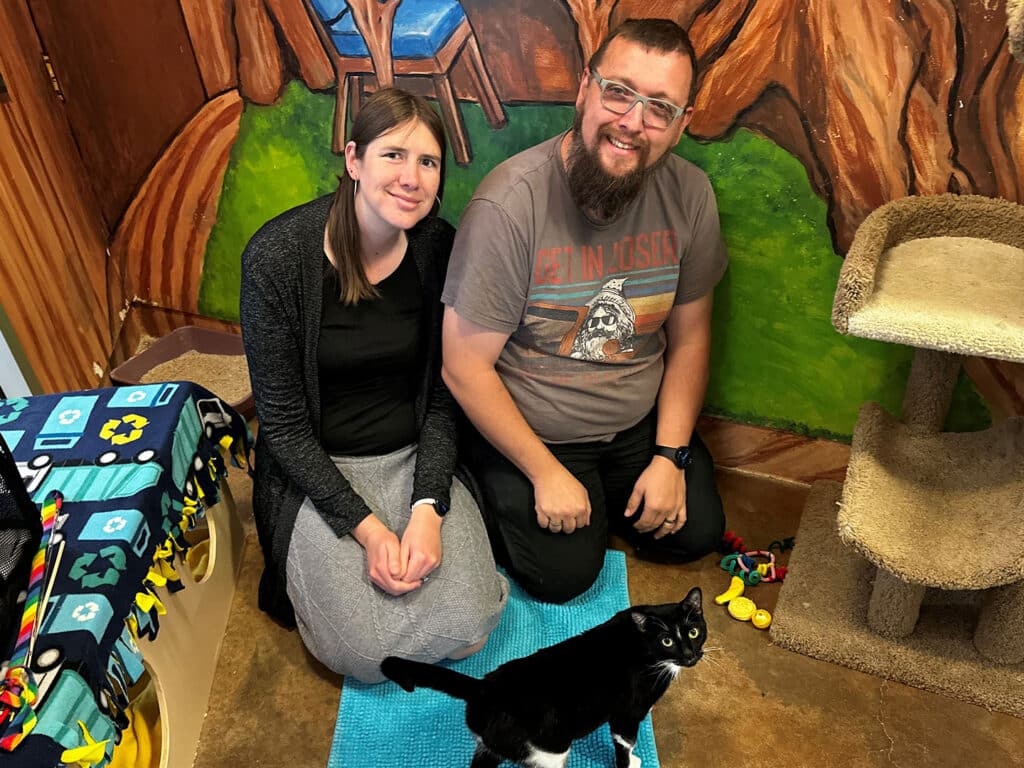 A woman and a man are sitting on a blue mat in a colorful animal shelter room, with a black and white cat nearby. The background features a mural of a tree and seating area, along with a cat tree and various colorful toys scattered around.