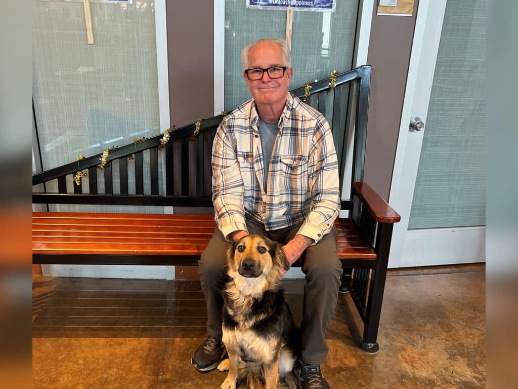 An older man with glasses wearing a plaid shirt and gray pants sits on a wooden bench with a black and brown dog beside him. The setting includes a modern interior with light-colored walls and a door in the background.