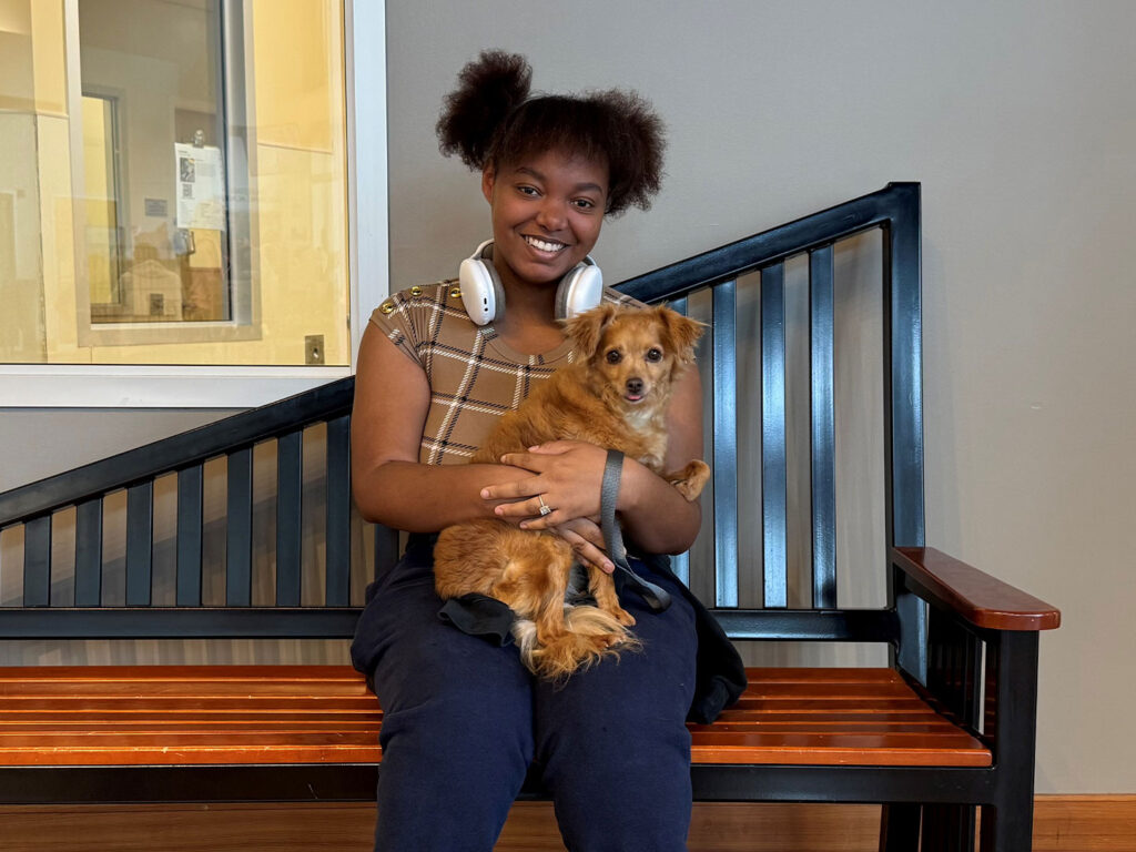 A young girl with curly hair smiles while sitting on a wooden bench, holding a small brown dog in her lap. She wears a plaid shirt and headphones, and a bright room is visible in the background.