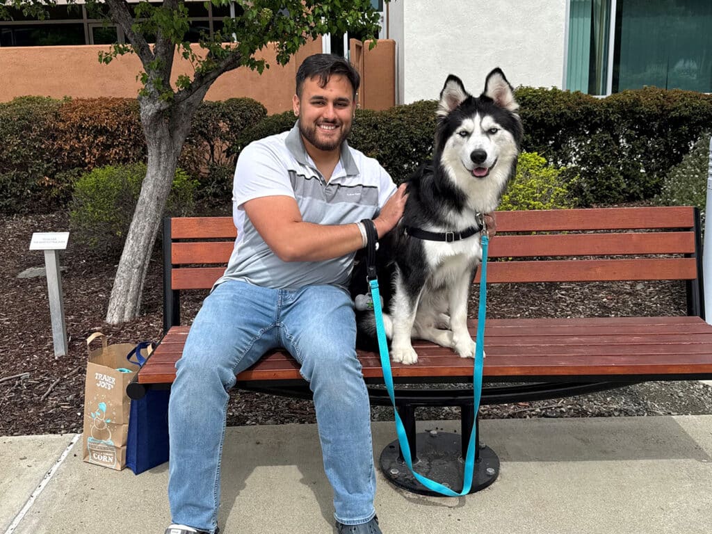 A man sitting on a bench with a black and white Siberian Husky dog beside him, both looking towards the camera. The man is smiling and wearing a gray shirt and jeans. A brown bag is placed next to the bench, and there are green bushes in the background.