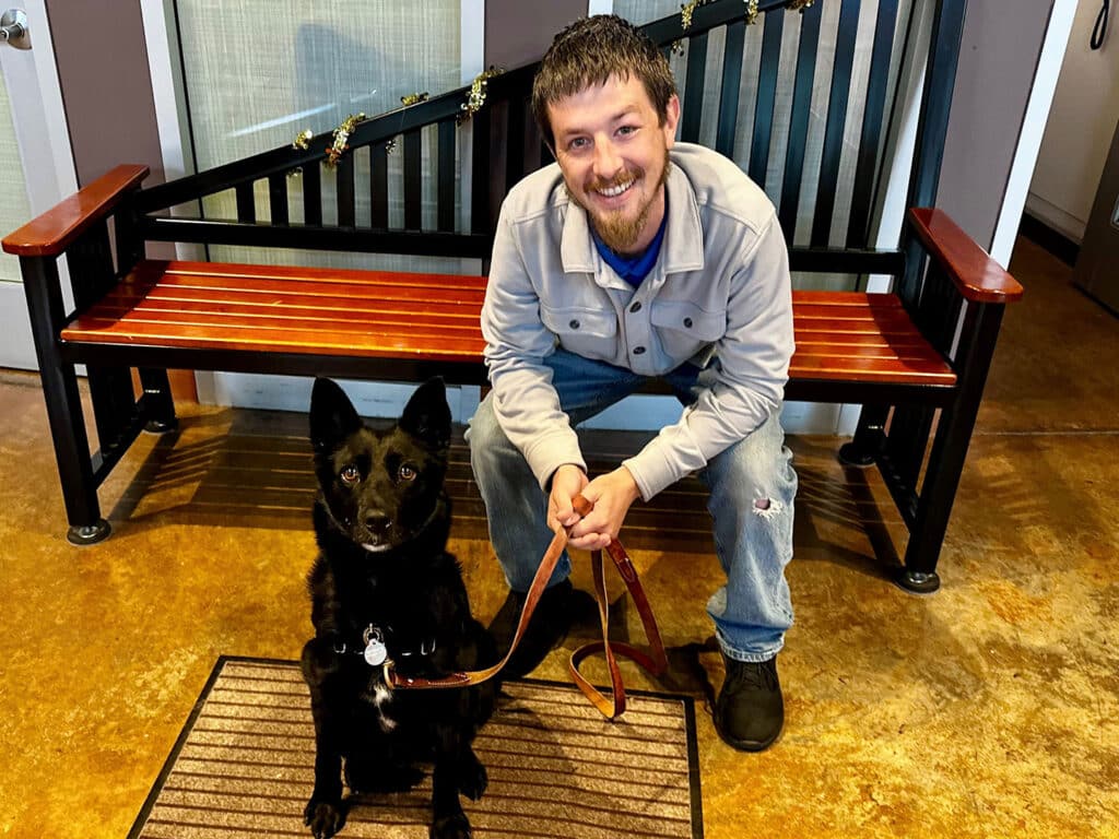 A man sitting on a wooden bench, smiling while holding a leash attached to a black dog. The dog sits in front of him on a welcome mat. The background shows a modern interior with light-colored walls and a decorative railing.