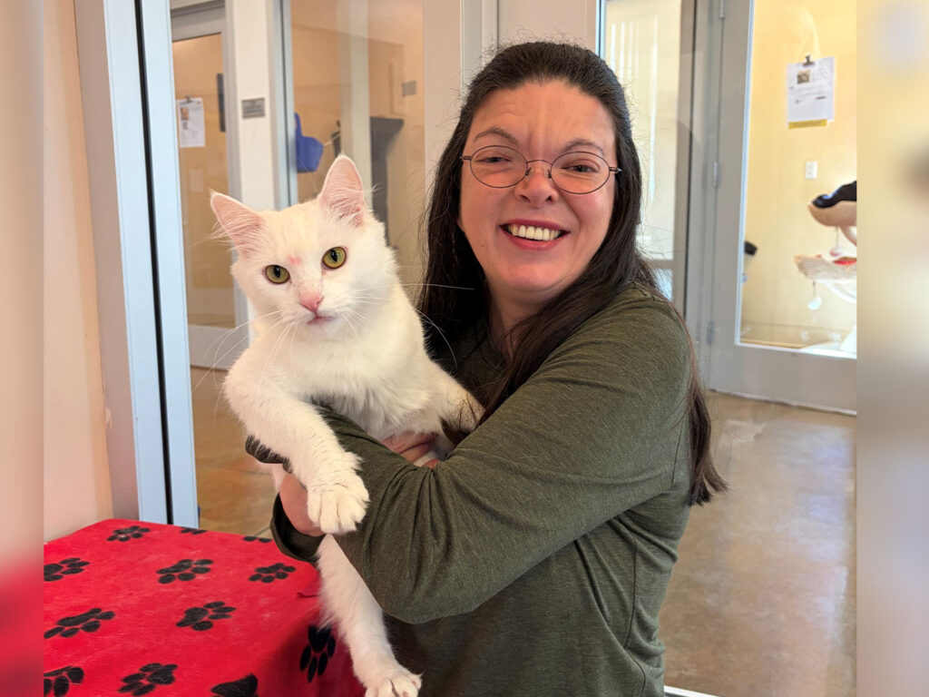 A woman with glasses smiles while holding a white cat in her arms. They are indoors with a red table covered in a black paw print pattern nearby, and glass doors visible in the background.