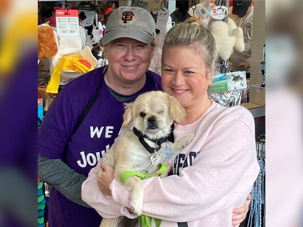 Two women smiling and holding a small dog in a pet store. The woman on the left wears a purple shirt and a gray cap, while the woman on the right wears a pink sweater. The dog, wearing a green harness, has light fur and a playful expression.