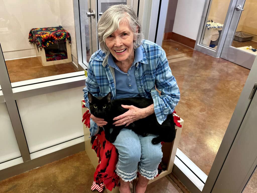 An elderly woman sitting in a chair holding a black cat on her lap. She is wearing a blue plaid shirt and light jeans, smiling at the camera. The setting is a bright animal adoption center with glass doors and a cozy atmosphere.