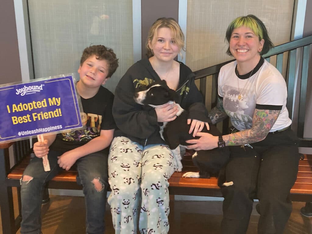 Two children and an adult sitting on a bench inside a facility. The boy holds a sign reading "I Adopted My Best Friend!" while the girl sits with a dog on her lap, and the woman has colorful hair and is placing her arm around the girl.